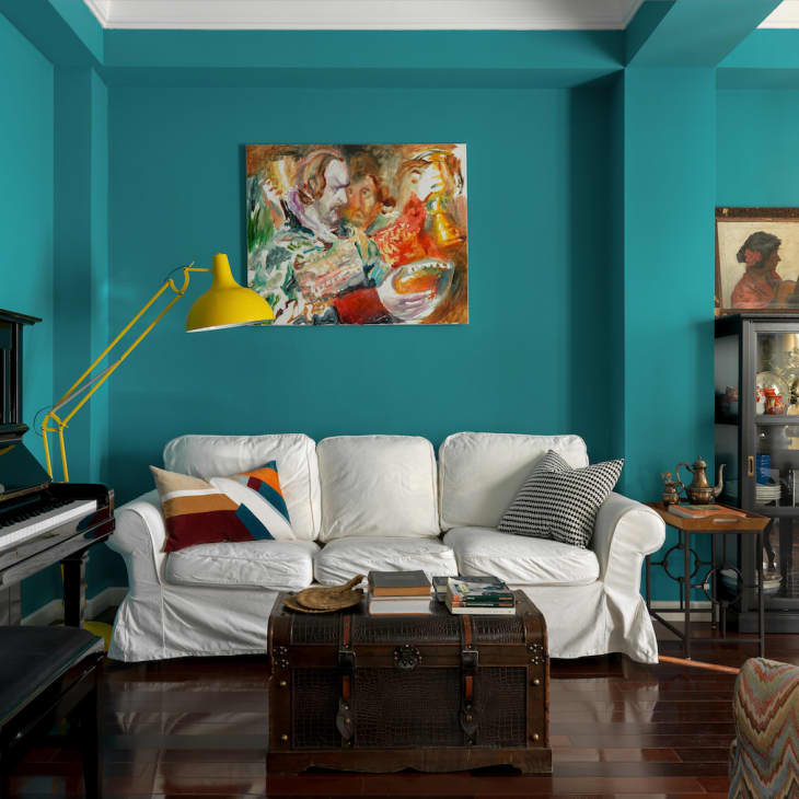 Teal living room with white sofa, colorful pillows, yellow lamp, piano, and eclectic artwork.