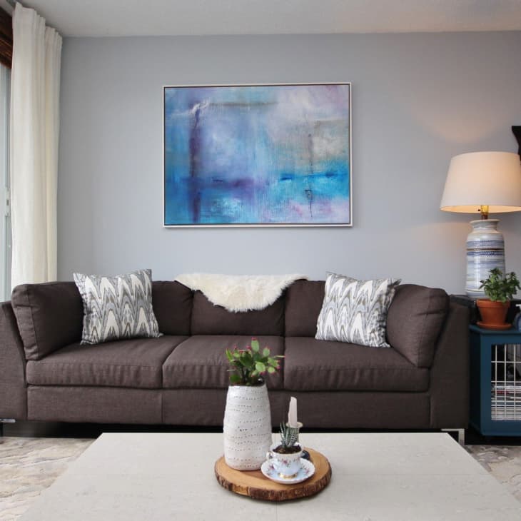 Gray sofa with patterned pillows, abstract blue artwork, and a lamp on a side table in a modern living room.