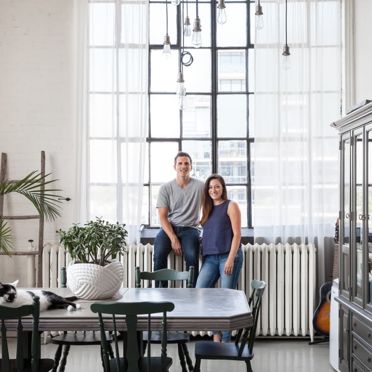 Couple standing in a bright dining room with a large window, green chairs, a cat on the table, and a globe on a cabinet.
