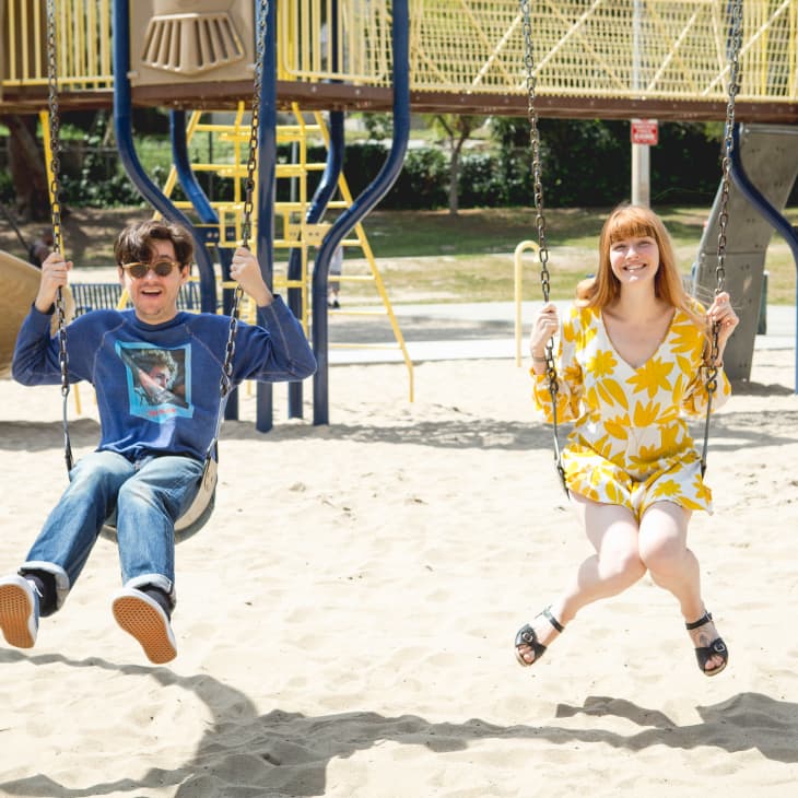 Two people swinging in a playground, one in a blue shirt and jeans, the other in a yellow floral dress.