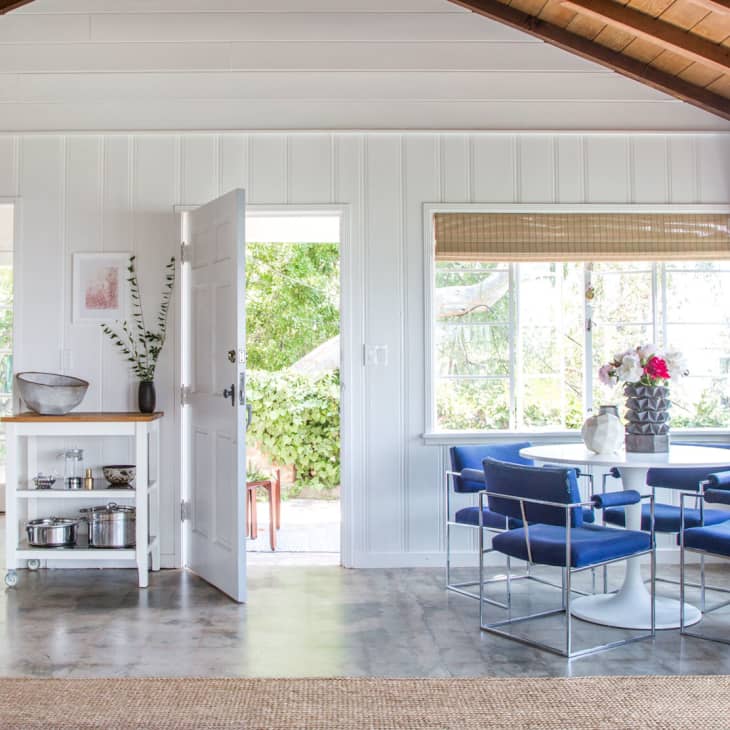 Open kitchen and dining area with blue chairs, round table, and large windows overlooking greenery.