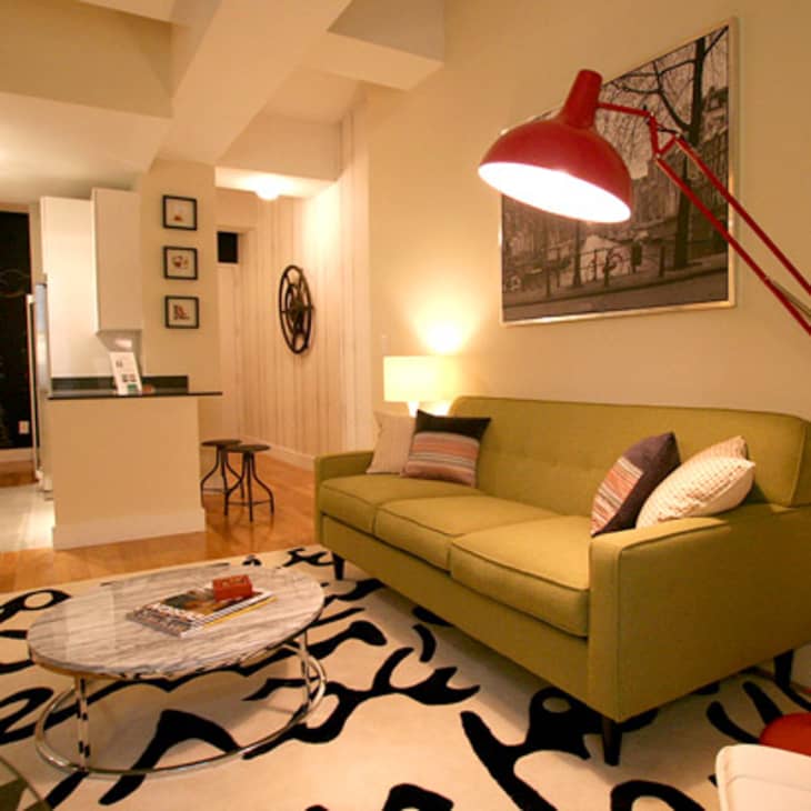 Living room with green sofa, red floor lamp, abstract rug, and wall art, leading to a kitchen with bar stools.