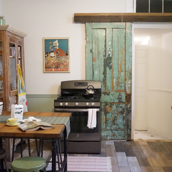 Rustic kitchen with vintage green door, wooden table, metal stools, and retro fridge. Brick wall and New Orleans poster.