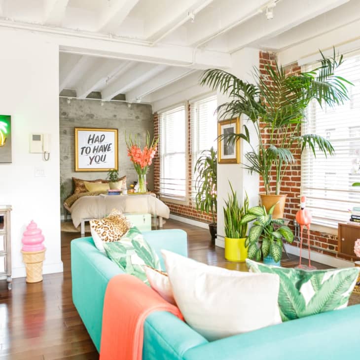 Bright living room with turquoise sofa, neon "Paradise" sign, tropical plants, and colorful decor. Bedroom visible in background.