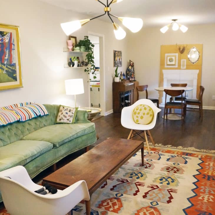 Living room with green sofa, colorful rug, modern chairs, and dining area in the background, featuring vibrant artwork and plants.