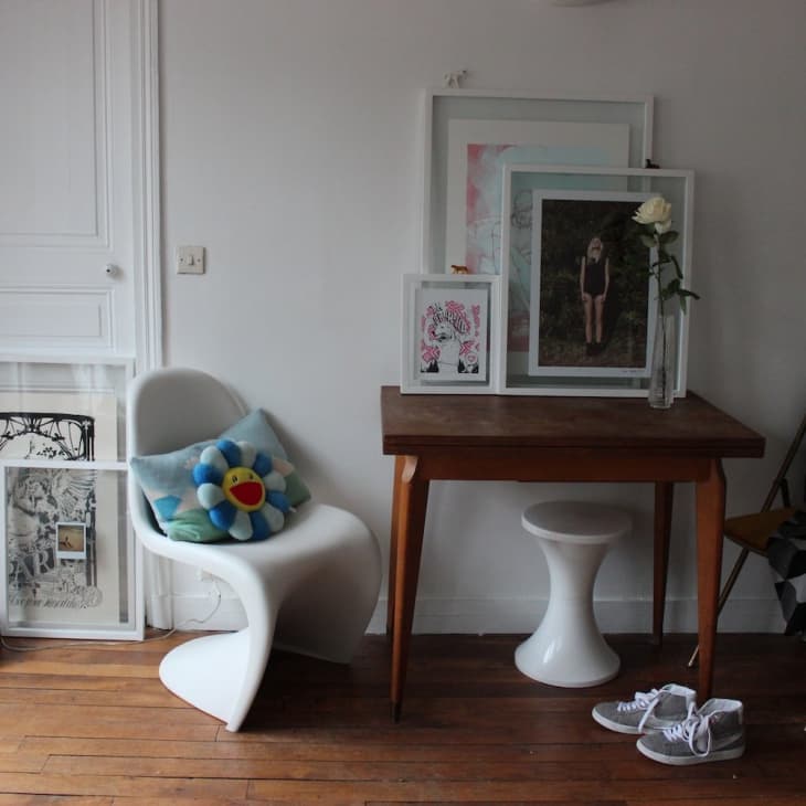 White chair with a flower pillow, wooden table with framed art, and a single rose in a vase on a wooden floor.