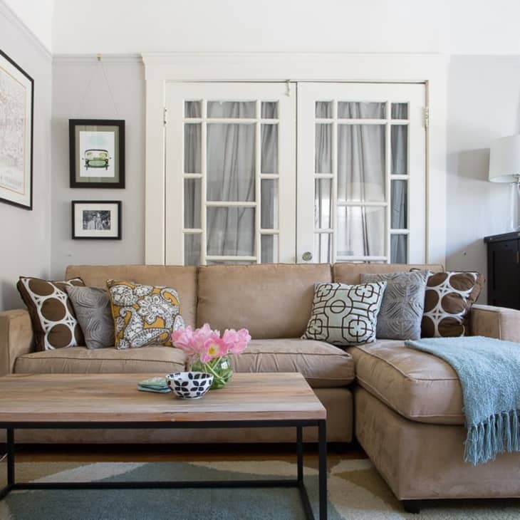 Beige sectional sofa with patterned pillows, wooden coffee table, and pink flowers in a living room with framed art and French doors.
