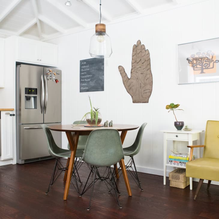 Kitchen with round wooden table, green chairs, stainless steel fridge, and yellow armchair. Wall art and potted plant decor.
