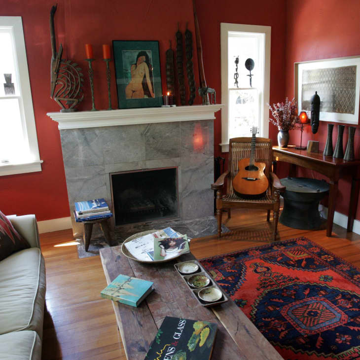 Cozy living room with red walls, a marble fireplace, guitar on a chair, and a vibrant patterned rug.
