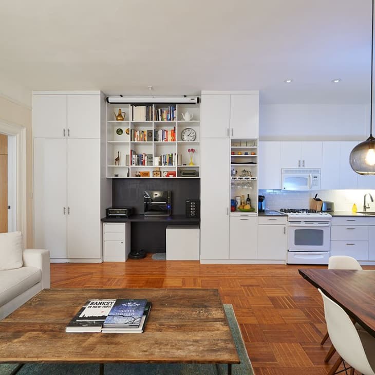 Open-plan living room and kitchen with white cabinets, wooden floors, a large sofa, dining table, and bookshelves.