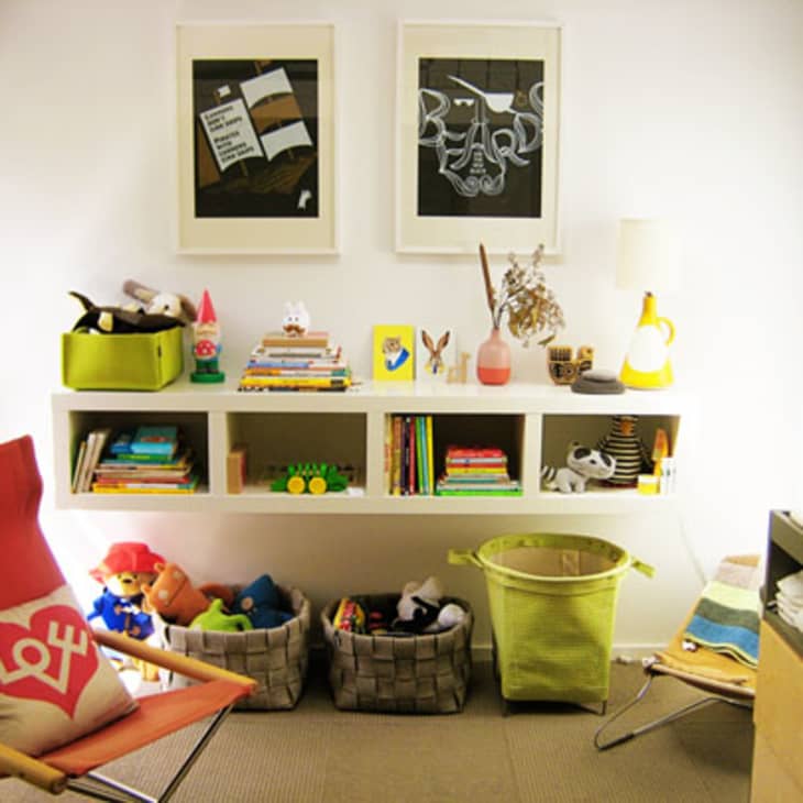 Children's room with wall shelves holding books and toys, a red chair with a "Love" pillow, and baskets on the floor.