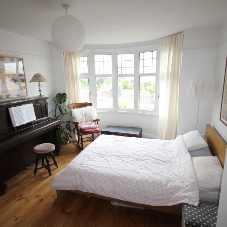 Bedroom with a piano, sheet music, wooden floor, and a bed near a large window with sheer curtains.