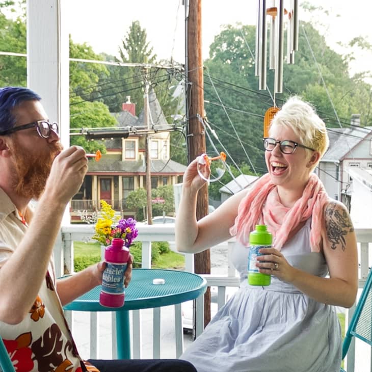 Two people blowing bubbles on a porch, sitting at a turquoise table with a view of houses and trees in the background.
