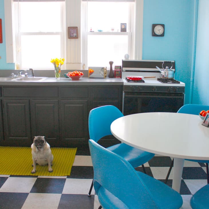 Retro kitchen with blue walls, black cabinets, checkered floor, round table, blue chairs, and a pug on a yellow rug.