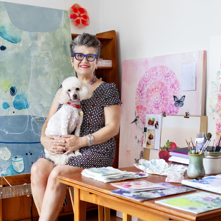 Artist with short hair and glasses holding a white poodle, seated in a studio with colorful abstract paintings and art supplies.