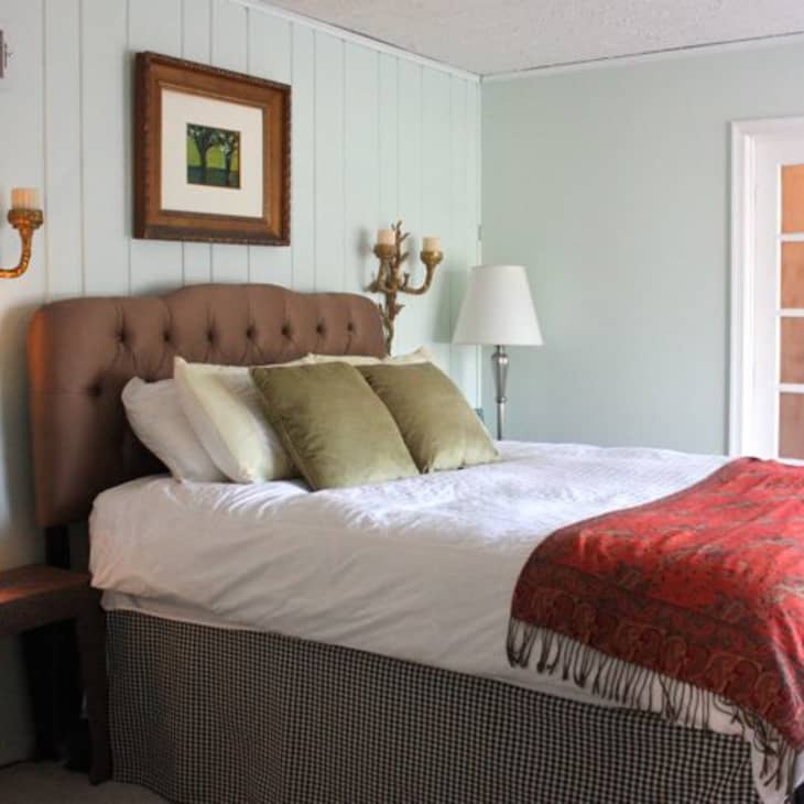Bedroom with tufted headboard, green pillows, red throw, wall sconces, framed art, and a side table with a lamp.