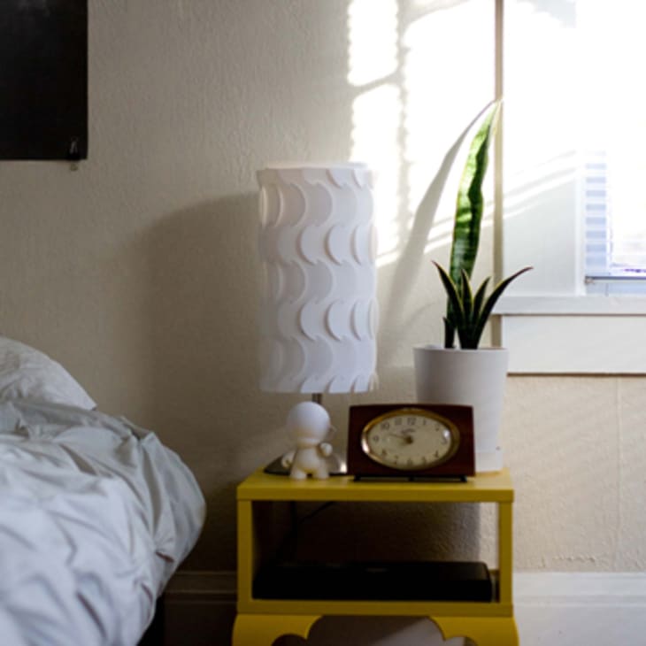 Bedroom with a yellow nightstand, white lamp, potted snake plant, and vintage clock beside a bed with white bedding.