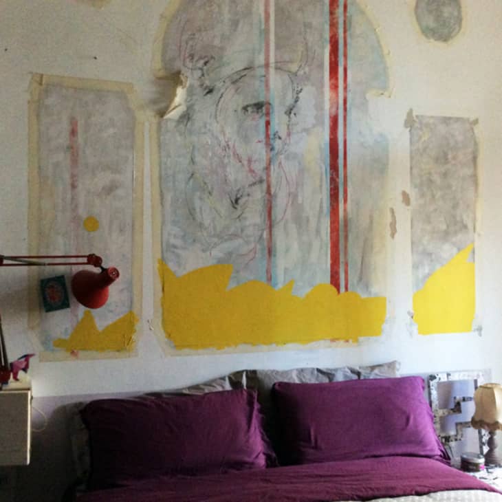 Bedroom with purple pillows, abstract wall art, red lamp, and a small bedside table with a lamp.