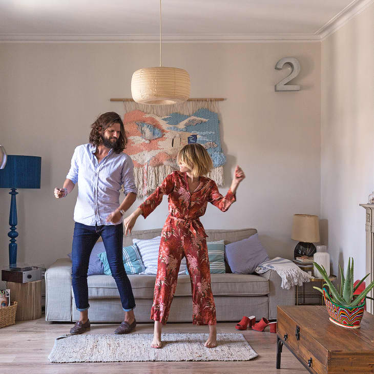 Couple dancing in a cozy living room with a gray sofa, colorful wall art, and a wooden coffee table with a potted plant.
