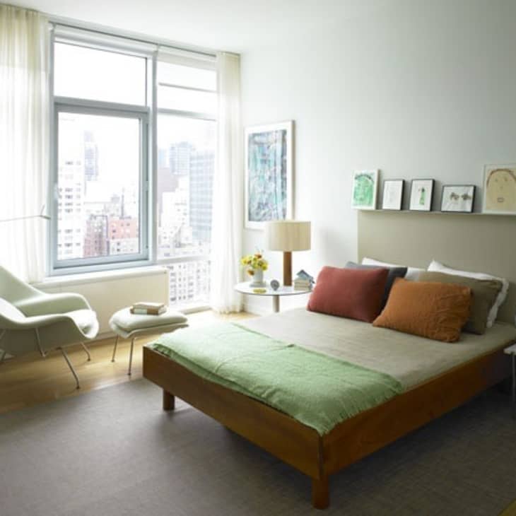 Modern bedroom with a wooden bed, green blanket, colorful pillows, and a city view through large windows.