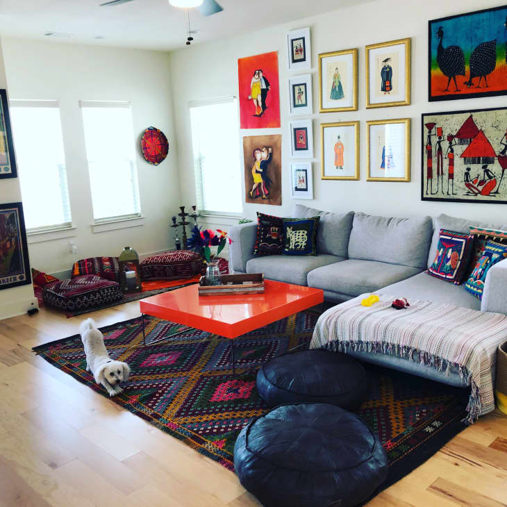 Living room with colorful art, gray sectional sofa, orange coffee table, patterned rugs, and a small white dog.