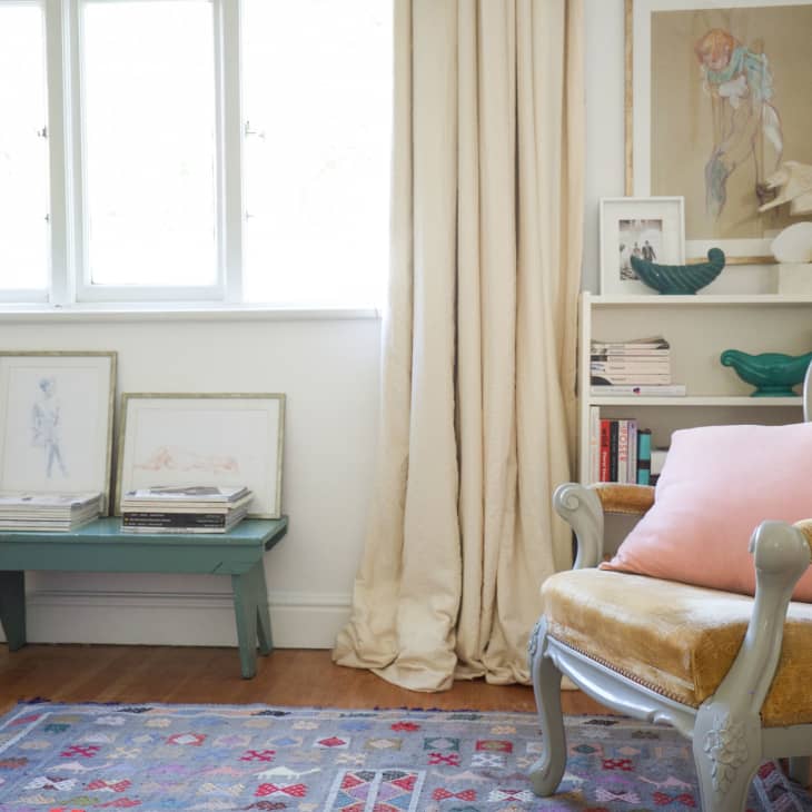 Cozy reading nook with a vintage armchair, pink pillow, books, and framed art on a shelf and table by a window.