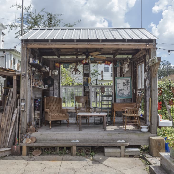 Rustic outdoor shed with vintage furniture, wooden chairs, and eclectic decor, surrounded by garden and string lights.