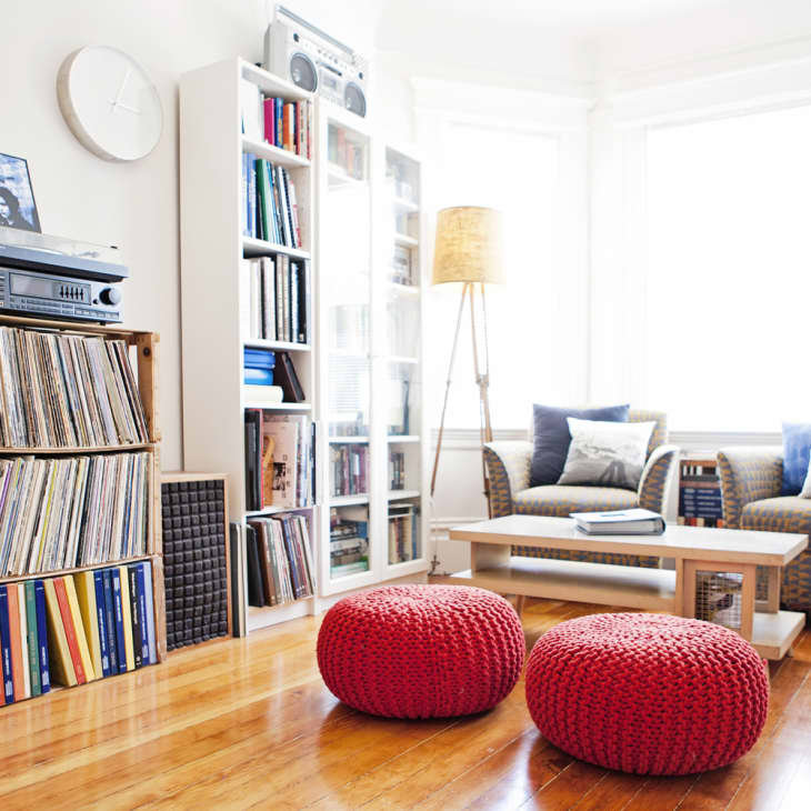 Cozy living room with wicker chairs, red poufs, wooden coffee table, and shelves filled with books and vinyl records.