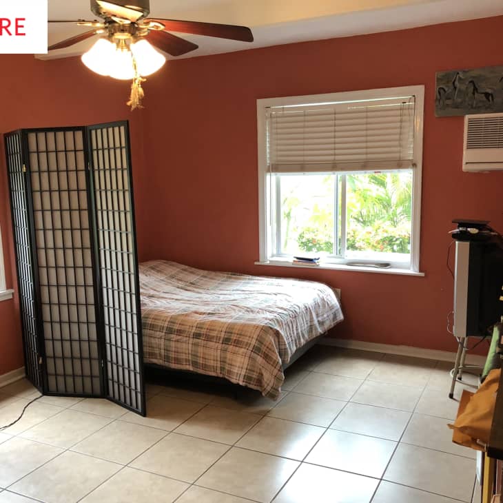 Bedroom with terracotta walls, bed with plaid bedding, folding screen, ceiling fan, and window AC unit.