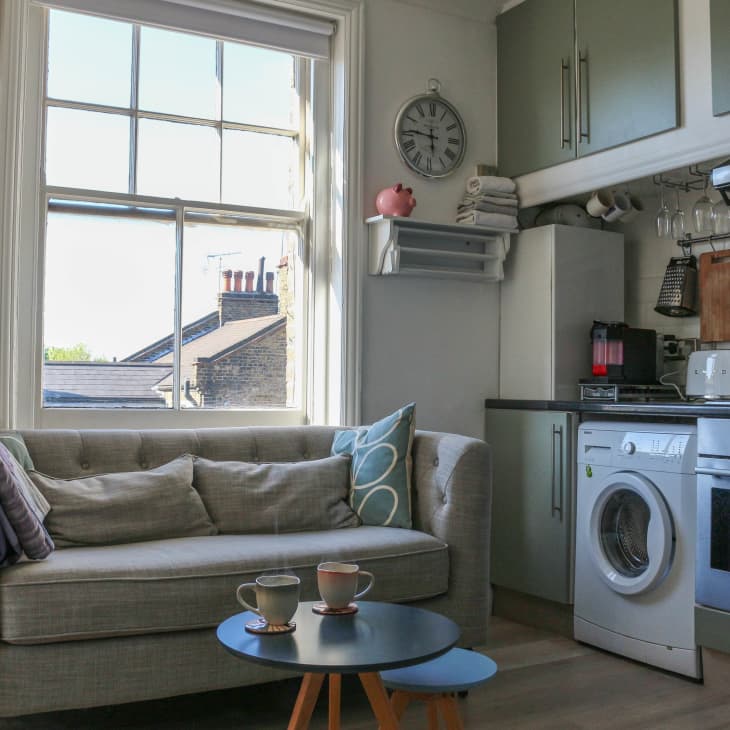 Cozy kitchen-living area with a gray sofa, wall art, washing machine, and kitchen appliances under green cabinets.