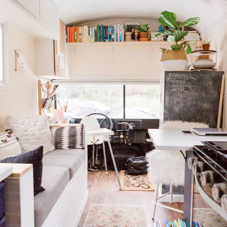 Cozy bus interior with a sofa, chalkboard, books, plants, and a small kitchen area featuring a stove and dining table.