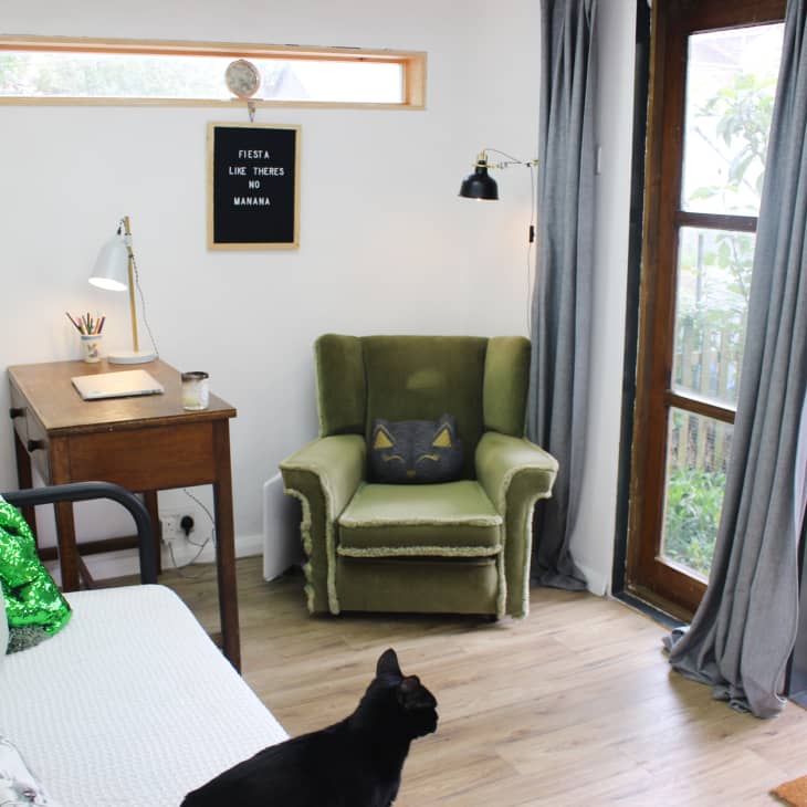 Cozy room with green armchair, wooden desk, laptop, and a black cat. Large windows with gray curtains and garden view.