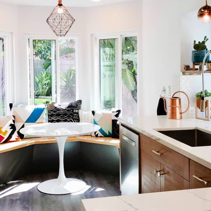 Bright kitchen nook with a round white table, colorful geometric pillows on a wooden bench, and potted plants on shelves.