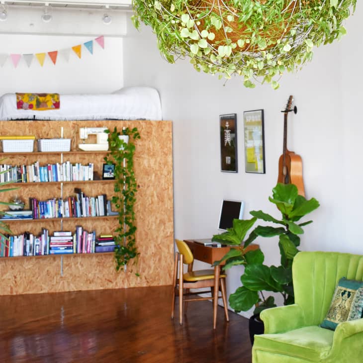 Loft with a bed above a bookshelf, green armchair, desk, guitar, and hanging plant.