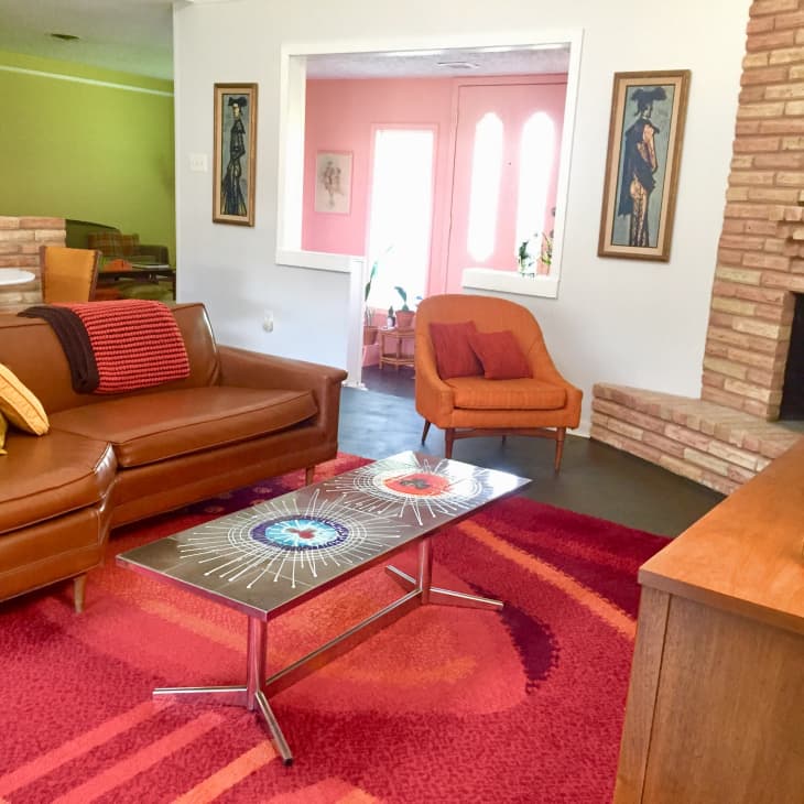 Mid-century living room with brown leather sofa, colorful cushions, orange armchair, geometric rug, and brick fireplace.