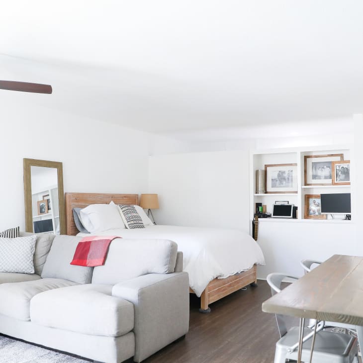 Open-plan studio with a gray sofa, wooden bed, red chimney, and wall-mounted TV.