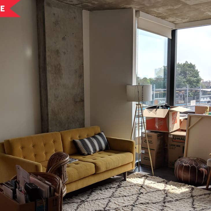Living room with yellow sofa, floor lamp, moving boxes, and large window with city view.