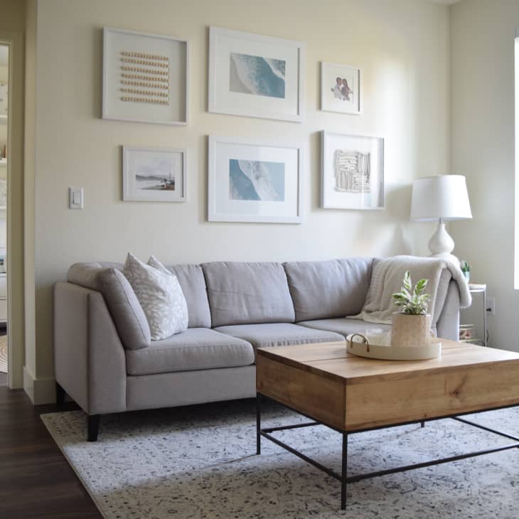 Living room with a gray sectional sofa, wooden coffee table, and framed art on the wall. Shelves with decor in the background.