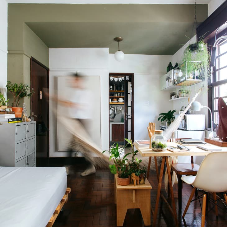 Cozy living room with plants, a wooden table, white chairs, and a bicycle by large windows. Person moving in the background.