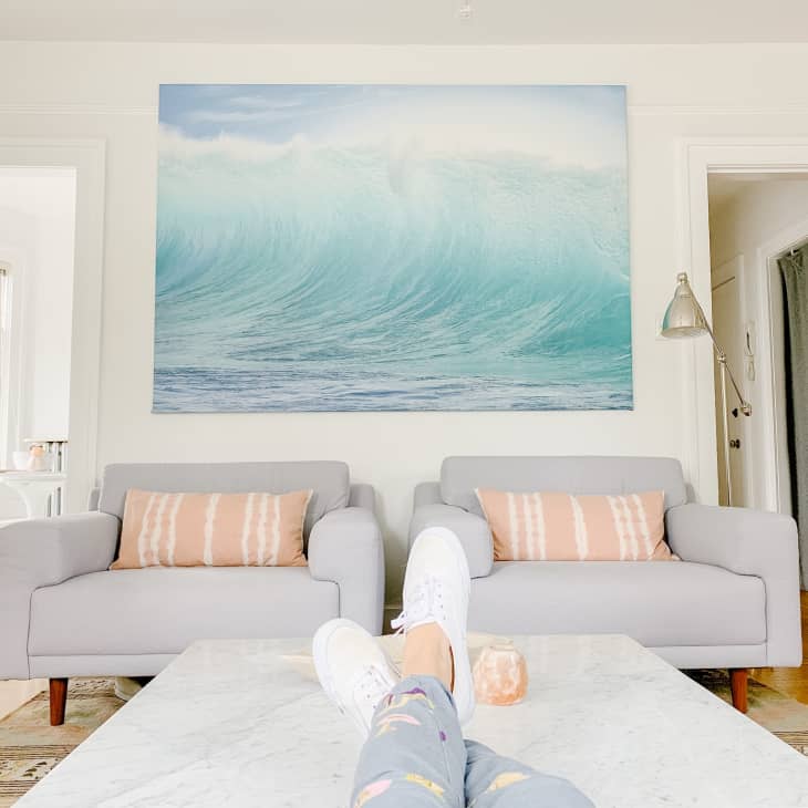 Person relaxing on a sofa with feet up on a marble table, facing a large ocean wave painting in a bright living room.
