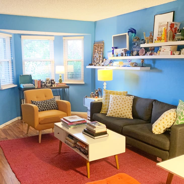 Cozy living room with blue walls, gray sofa, yellow armchair, colorful pillows, and shelves with books and decor.