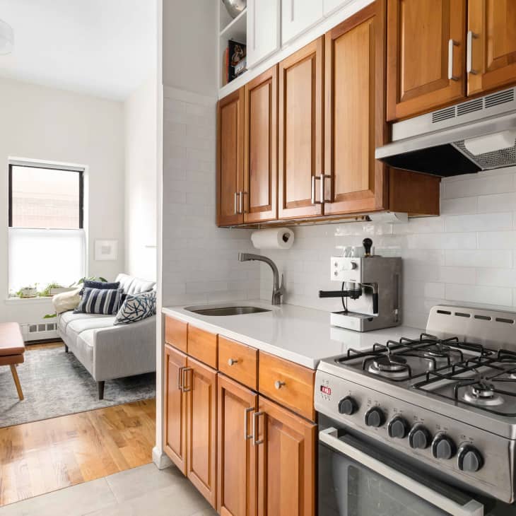Compact kitchen with wooden cabinets, gas stove, espresso machine, and adjacent living area with a gray sofa and window.