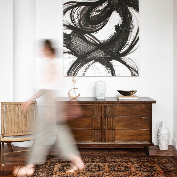 Blurred person walking past a wooden sideboard with abstract art, wicker chair, and decorative items in a modern room.