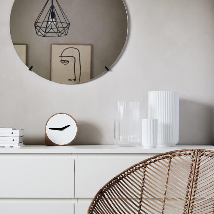 Round mirror above white dresser with books, clock, and vases; wicker chair in foreground.