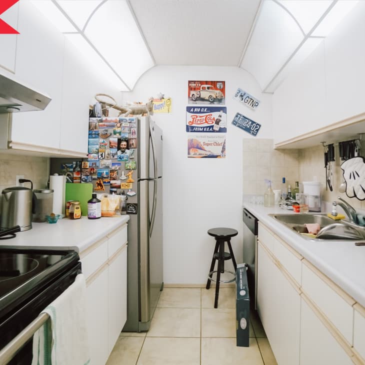 Narrow kitchen with white cabinets, stainless steel fridge covered in magnets, and a black stool.