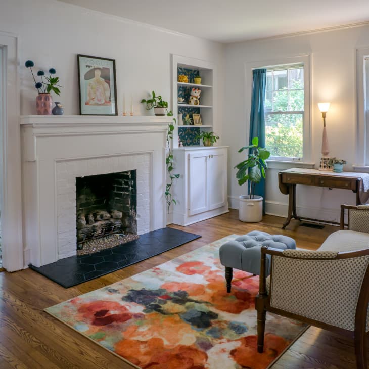 Living room with a white fireplace, colorful rug, blue curtains, armchair, and potted plants by the window.