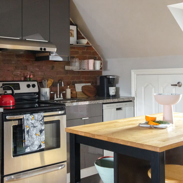 Modern kitchen with gray cabinets, brick backsplash, stainless steel stove, red kettle, and wooden island with fruit bowl.