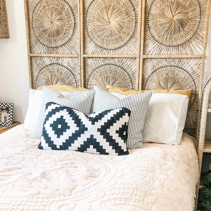 Bohemian bedroom with woven headboard, patterned pillows, wooden side table, and decorative shelf with plants and pottery.