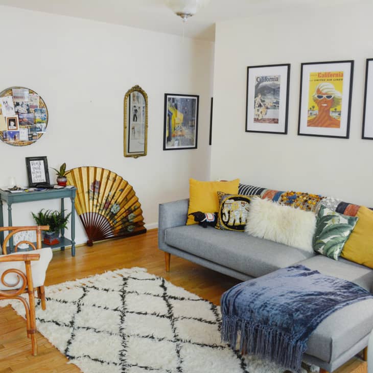 Cozy living room with gray sectional sofa, colorful pillows, rattan chair, wall art, and decorative fan on wooden floor.