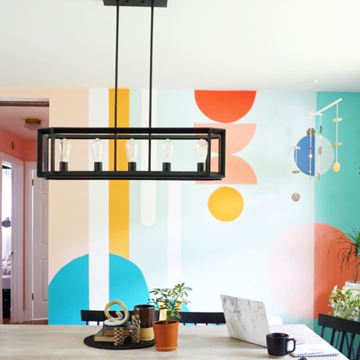 Dining room with colorful geometric wall mural, black chandelier, plants, and a table with a laptop and mug.